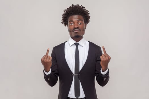 Impolite displeased man with Afro hairstyle shows middle finger, being angry with someone, purses lips frowns furious, wearing white shirt and tuxedo. Indoor studio shot isolated on gray background.