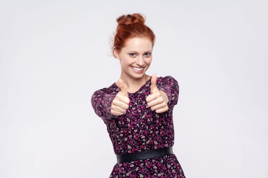 Portrait of smiling happy satisfied ginger woman wearing dress standing showing thumbs up, recommend excellent service. Indoor studio shot isolated on gray background.