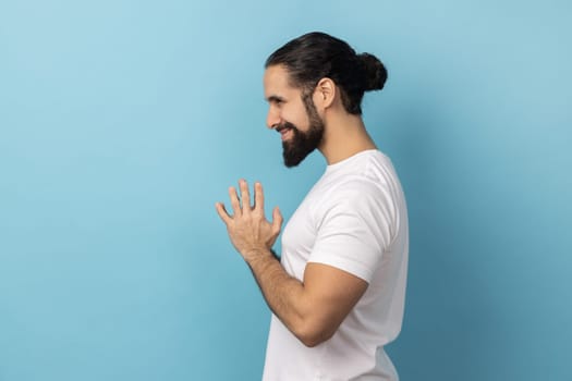 Side view of devious man with beard wearing white T-shirt smirking and conspiring cunning sly plan, pondering tricky clever idea in mind, wants to cheat. Indoor studio shot isolated on blue background