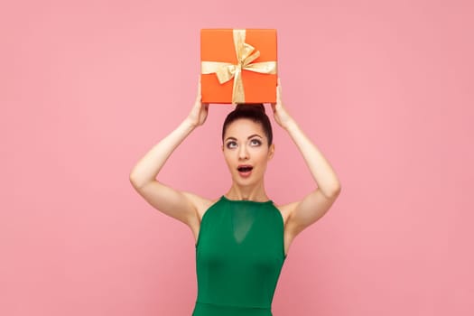 Portrait of surprised astonished amazed woman standing holding red present box above her head, being excited, wearing green dress. Indoor studio shot isolated on pink background.