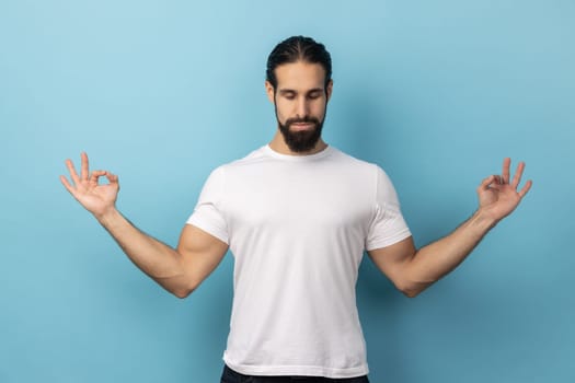 Portrait of calm relaxed man with beard wearing white T-shirt doing yoga, breathing, keeping eyes closed, calming down after work. Indoor studio shot isolated on blue background.