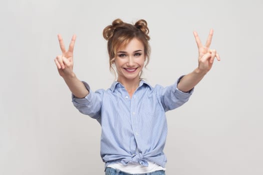 Portrait of joyful blonde woman showing peace v sign, smiling toothily, enjoying nice day, gestures victory symbol, wearing blue shirt. Indoor studio shot isolated on gray background.