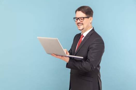 Portrait of smiling cheerful man standing and holding notebook, working on laptop online and joying his work, wearing black suit with red tie. Indoor studio shot isolated on light blue background.