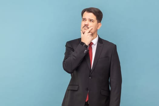 Portrait of pensive thoughtful man with mustache standing with puzzled expression, looking away, holding chin, wearing black suit with red tie. Indoor studio shot isolated on light blue background.