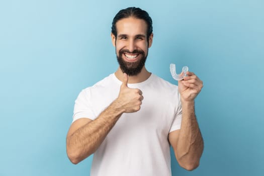 Portrait of man with beard wearing white T-shirt holding dental aligner retainer, dental clinic for beautiful teeth, showing thumb up. Indoor studio shot isolated on blue background.