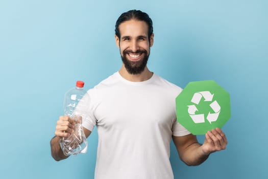 Portrait of responsible smiling man with beard wearing white T-shirt holding empty plastic bottle and green recycling sign, looking at camera. Indoor studio shot isolated on blue background.