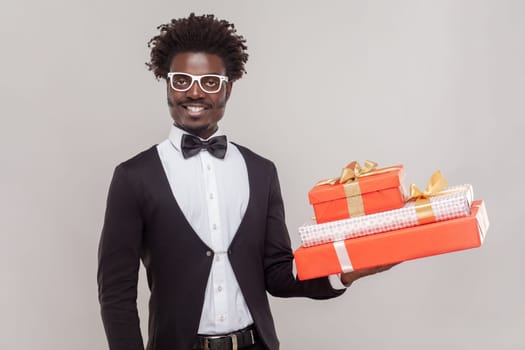 Portrait of joyful cheerful man in glasses standing holding three present boxes, being in festive mood, wearing white shirt and tuxedo. Indoor studio shot isolated on gray background.