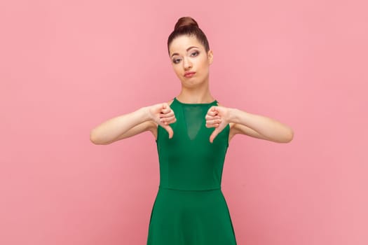 Portrait of beautiful disappointed woman with bun hairstyle standing showing thumb down, looks unhappy, dislikes service, wearing green dress. Indoor studio shot isolated on pink background.