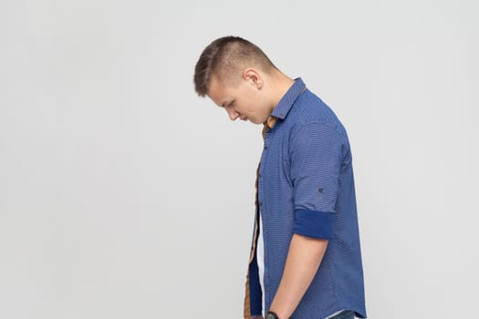 Side view portrait of upset teenager boy wearing blue shirt standing with his head down and looking, having with dissatisfied sadness face. Indoor studio shot isolated on gray background.