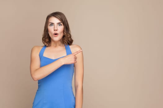 Portrait of surprised woman with wavy hair showing something aside at blank wall, being astonished, advertisement area, wearing blue dress. Indoor studio shot isolated on light brown background.