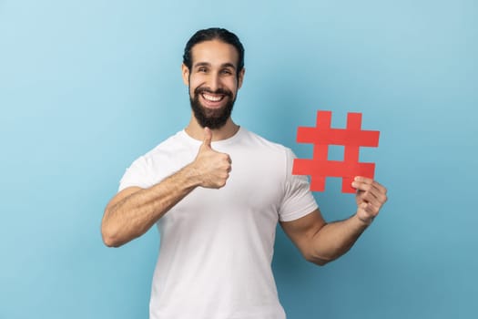 Portrait of happy man with beard wearing white T-shirt holding hashtag symbol, showing thumb up, recommending popular topics, internet trends. Indoor studio shot isolated on blue background.