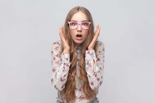 Portrait of astonished shocked woman in glasses with long blond hair looking at camera, keeping mouth widely open, keeps hands raised. Indoor studio shot isolated on gray background.