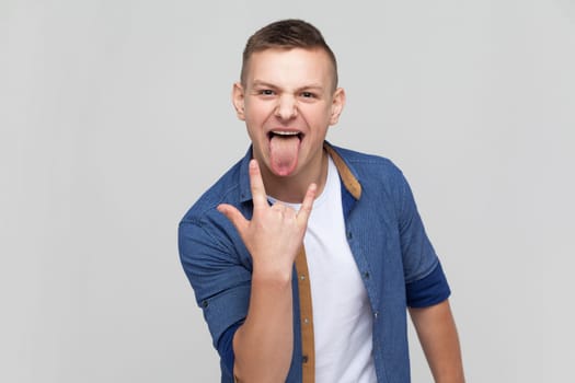 Portrait of enthusiastic crazy teenager boy in blue shirt showing rock and roll gesture and sticking out tongue, feeling to be cool rocker on festival. Indoor studio shot isolated on gray background.