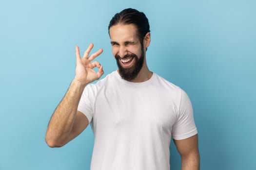 Portrait of satisfied happy man with beard wearing white T-shirt standing, looking at camera showing Ok sign gesture, expressing positive emotions. Indoor studio shot isolated on blue background.