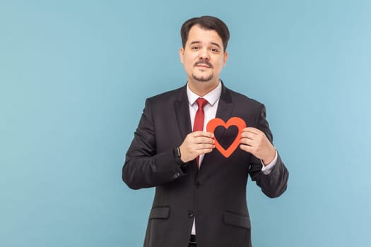 Portrait of attractive man with mustache standing holding red gesture, looking at camera with positive expression, wearing black suit with red tie. Indoor studio shot isolated on light blue background