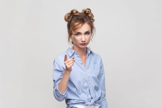 Hey, listen me attentively. Portrait of blonde woman raising index finger, looking with angry expression, warning you about danger, wearing blue shirt. Indoor studio shot isolated on gray background.