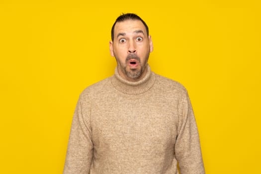 Bearded hispanic man in his 40s wearing a beige turtleneck tremendously amazed and surprised with open mouth in shock, isolated over yellow background