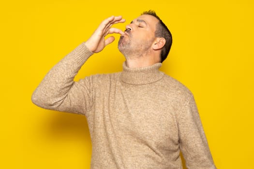 Bearded Hispanic man wearing a beige turtleneck smacking his lips and licking his fingers after devouring an exquisite delicacy, isolated on yellow studio background