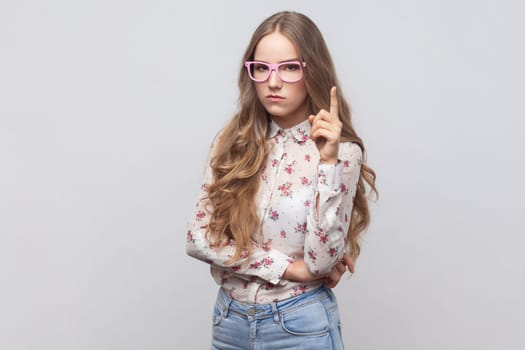 Be careful. Portrait of beautiful woman in glasses with long blond hair seriously pointing finger up and warning, looking at camera. Indoor studio shot isolated on gray background.