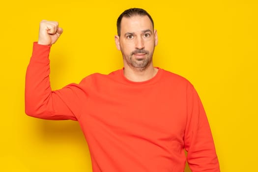 Bearded hispanic man wearing a red jersey flexing his biceps muscle showing his pitiful state of shape, isolated over yellow background