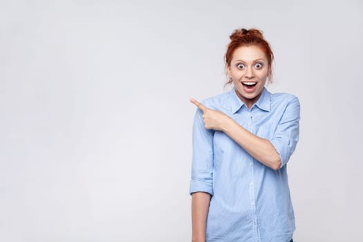 Portrait of shocked ginger woman wearing blue shirt looking at camera and pointing away, presenting copy space for advertisement or promotion. Indoor studio shot isolated on gray background.