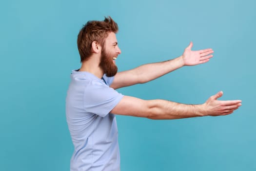 Free hugs, come into my arms. Side view of happy bearded man stretching hands to camera and smiling broadly, going to embrace, share love. Indoor studio shot isolated on blue background.