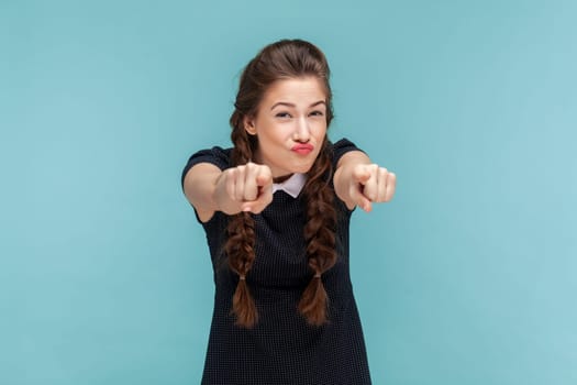 Portrait of funny attractive young woman with braids pointing at you, looking at camera with frowning face, wearing black dress. woman Indoor studio shot isolated on blue background.