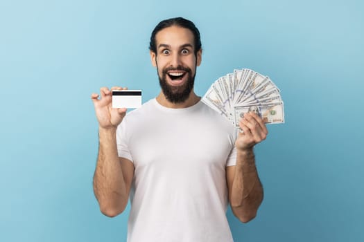 Amazed man wearing white T-shirt holding credit card and dollar banknotes, feeling rich with lot of money, excited to get bank loan for shopping. Indoor studio shot isolated on blue background.