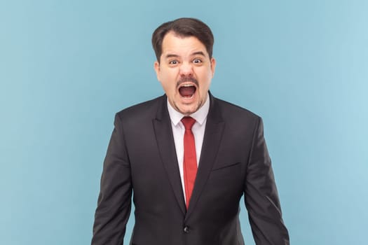 Portrait of angry furious man with mustache standing standing and screaming loud with hate and anger, wearing black suit with red tie. Indoor studio shot isolated on light blue background.