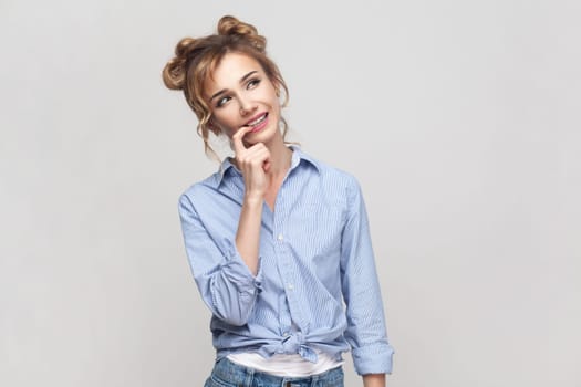 Portrait of pleased dreamy young adult blonde woman looks away with dreamy expression dreams about something, wearing blue shirt. Indoor studio shot isolated on gray background.