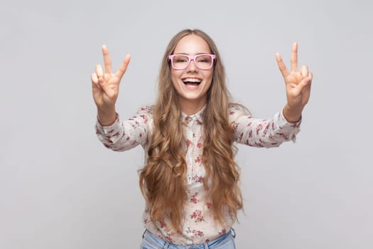 Belief in winning. Smiling pretty woman in glasses with wavy blond hair showing v sign with fingers, peace or victory gesture, rejoicing success. Indoor studio shot isolated on gray background.