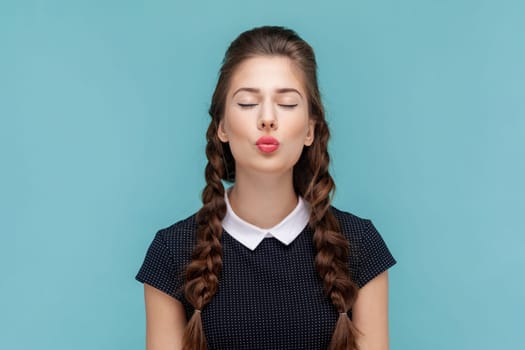 Portrait of romantic flirting woman with braids standing with closed eyes and pout lips, sending air kisses to her boyfriend, wearing black dress. woman Indoor studio shot isolated on blue background.