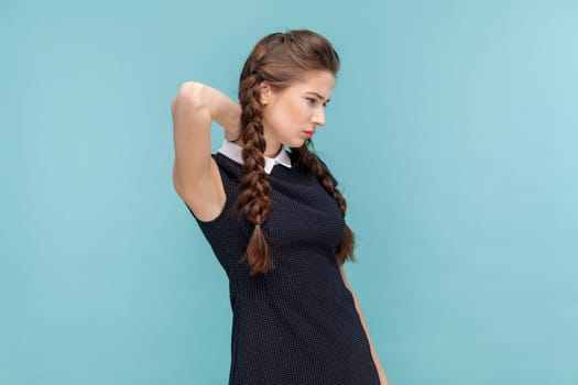 Portrait of unhappy sick woman with braids feels pain in neck, grimaces from pain, suffers without painkillers, wearing black dress. woman Indoor studio shot isolated on blue background.