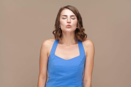 Portrait of flirting romantic beautiful woman with wavy hair standing with closed eyes and sending air kissing, wearing blue dress. Indoor studio shot isolated on light brown background.