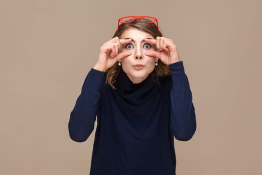 Portrait of funny surprised astonished woman with wavy hair in red red glasses on her head opens her eyes with fingers to see better. Indoor studio shot isolated on light brown background.