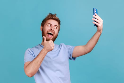 Portrait of positive smiling bearded man showing thumbs up to smartphone, communicating by video call, taking selfie and gesturing like. Indoor studio shot isolated on blue background.