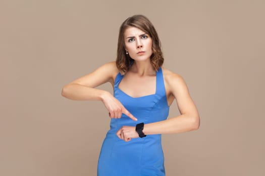 You are late. Portrait of serious woman with wavy hair standing, looking at camera and pointing at her smart watch, wearing blue dress. Indoor studio shot isolated on light brown background.