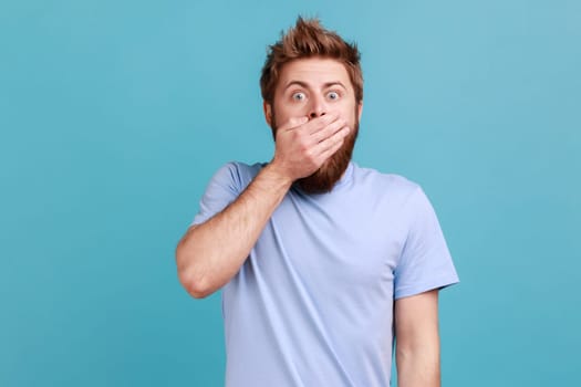 Portrait of shocked astonished bearded man standing covering his mouth and looking at camera with unbelievable face, keeping secret information. Indoor studio shot isolated on blue background.