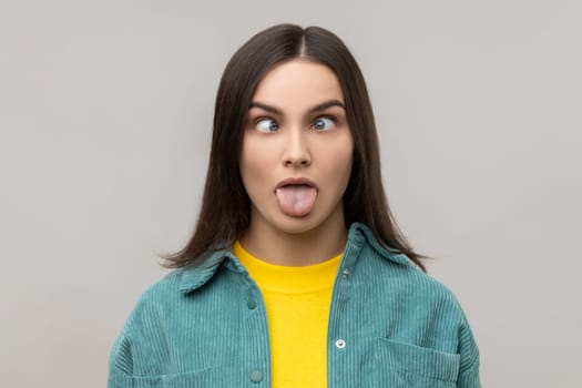 Portrait of dark haired woman sticking out tongue and looking at camera with crossed eyes, teasing with naughty expression, disobedient behavior. Indoor studio shot isolated on gray background.