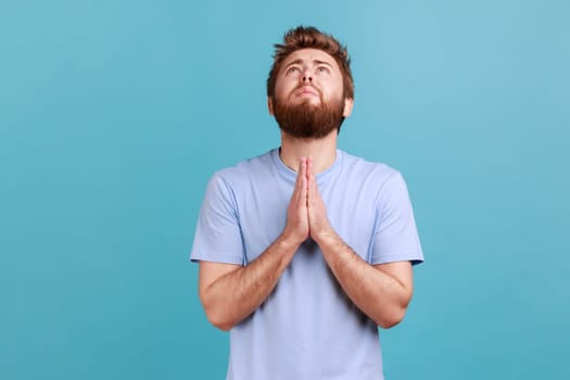 Portrait of hopeless desperate bearded man folding hands in pray looking up, communicating with god, asking for forgiveness and blessing. Indoor studio shot isolated on blue background.