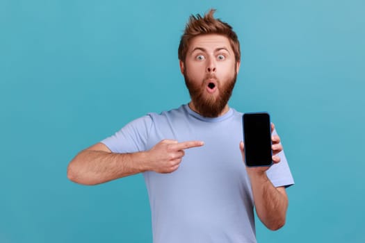 Portrait of astonished shocked bearded man pointing finger at smartphone with black blank screen, copy space for advertisement or promotional text. Indoor studio shot isolated on blue background.