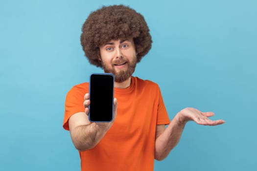 Portrait of confused man with Afro hairstyle wearing orange T-shirt showing cell phone with blank black screen, presenting area for advertisement. Indoor studio shot isolated on blue background.