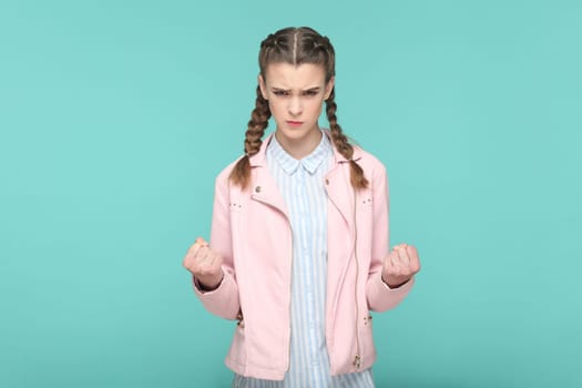 Portrait of angry aggressive attractive teenager girl with braids wearing pink jacket standing with clenched fits, arguing with somebody. Indoor studio shot isolated on green background.