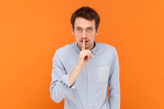 Serious young adult man standing with finger near lips, asking to keep silent, looking at camera with bossy expression, wearing light blue shirt. Indoor studio shot isolated on orange background.