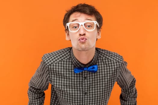 Portrait of man nerd keeps lips folded, expressing love, blows mwah, has romantic expression, wearing shirt with blue bow tie and white glasses. Indoor studio shot isolated on orange background.