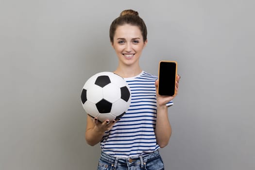 Portrait of woman wearing striped T-shirt looking at camera with toothy smile, holding soccer ball, showing cell phone with black blank screen. Indoor studio shot isolated on gray background.