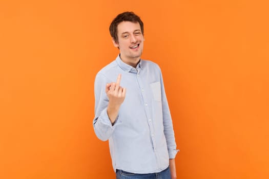 Portrait of serious vulgar selfish man showing middle finger, impolite gesture, disrespects someone, looks rude at camera, wearing light blue shirt. Indoor studio shot isolated on orange background.