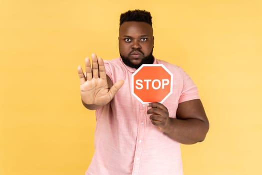 Portrait of man wearing pink shirt holding red stop sign, looking at camera with ban palm gesture, has strict expression, prohibition. Indoor studio shot isolated on yellow background.