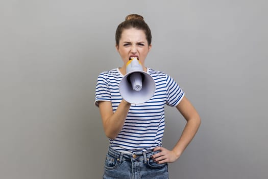 Portrait of woman in striped T-shirt holding megaphone near mouth loudly speaking, screaming, making announcement, paying attention at social problems. Indoor studio shot isolated on gray background.