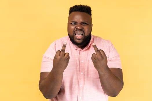 Portrait of impolite aggressive man wearing pink shirt showing middle fingers and asking to get off expressing negativity, disrespectful behaviour. Indoor studio shot isolated on yellow background.